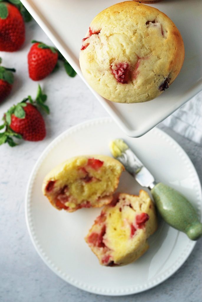 Close up of a strawberry muffin with fresh strawberries and a muffin sliced in half and buttered in the backgroun