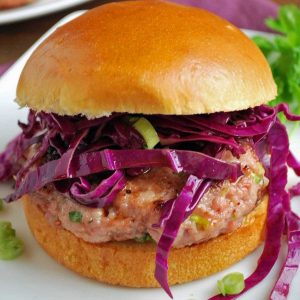 juicy pork burger patty topped with shredded red cabbage and sliced green onions on a brioche bun on a white plate