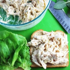 Open-faced chicken salad sandwich on wheat bread with a knife, fresh sage leaves, and a bowl of chicken salad in the background