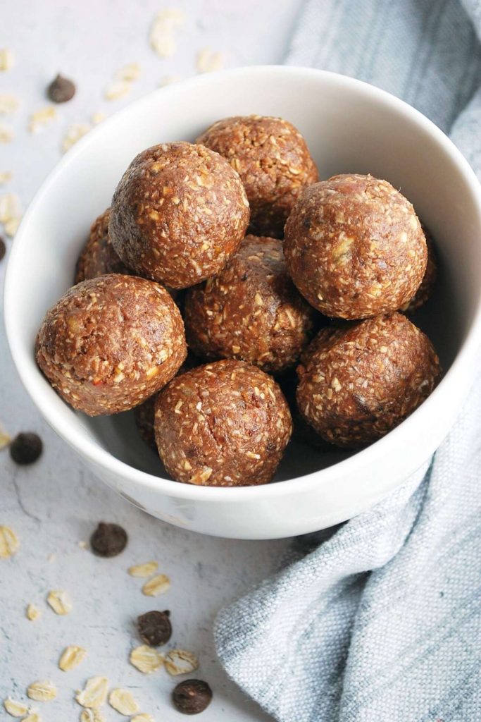 overhead photo of chocolate protein date balls in a white bowl with a napkin, chocolate chips and oats around the bowl.