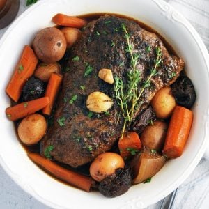 overhead photo of pot roast in a white serving dish with carrots, potatoes, and fresh thyme
