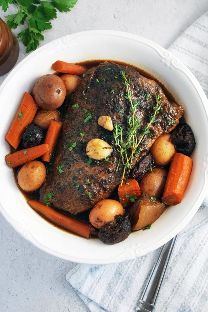 A white serving dish containing pot roast with carrots, potatoes, and prunes with a sprig of fresh thyme