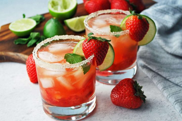 side view of two strawberry mojitos with a sugared rim and fresh lime and strawberries