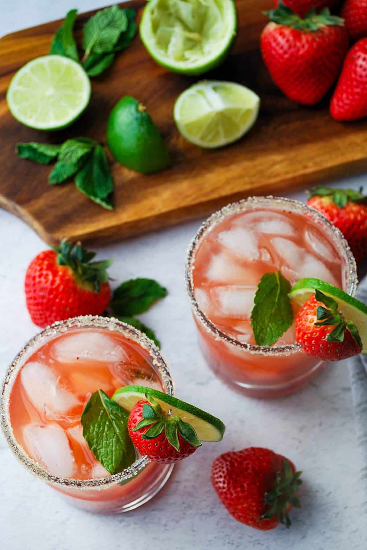 looking down at two strawberry cocktails with a cutting board with limes, strawberries, and fresh mint on it