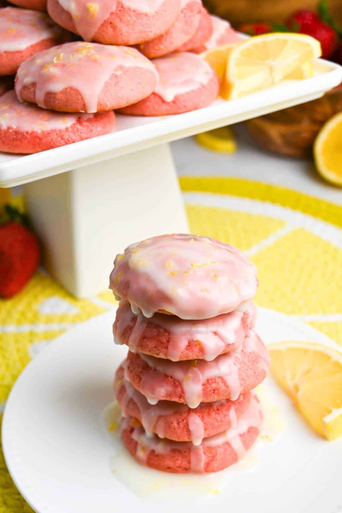 a stack of strawberry cookies dripping with icing and a platter of cookies behind it