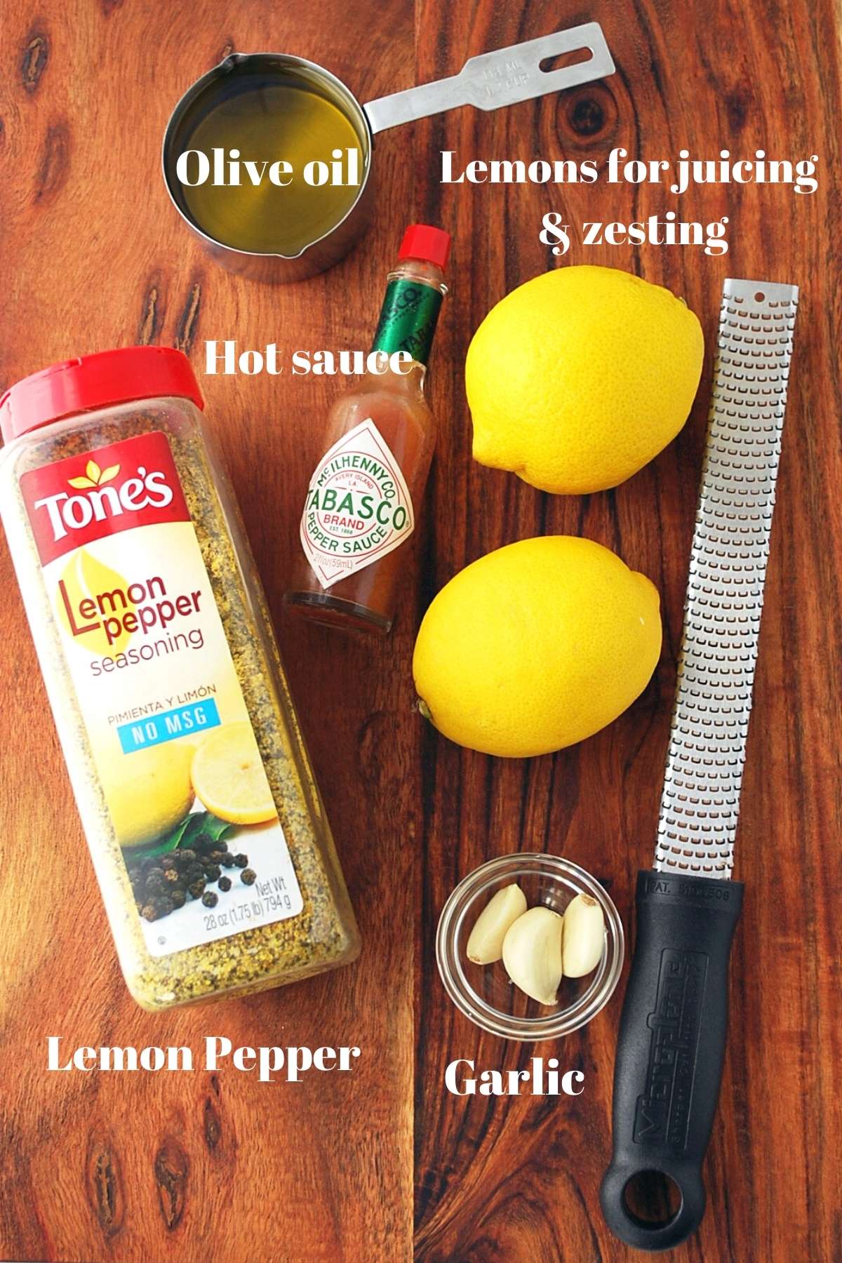 olive oil, lemon pepper, Tabasco, 2 lemons, garlic cloves, and a Microplane zester on a cutting board