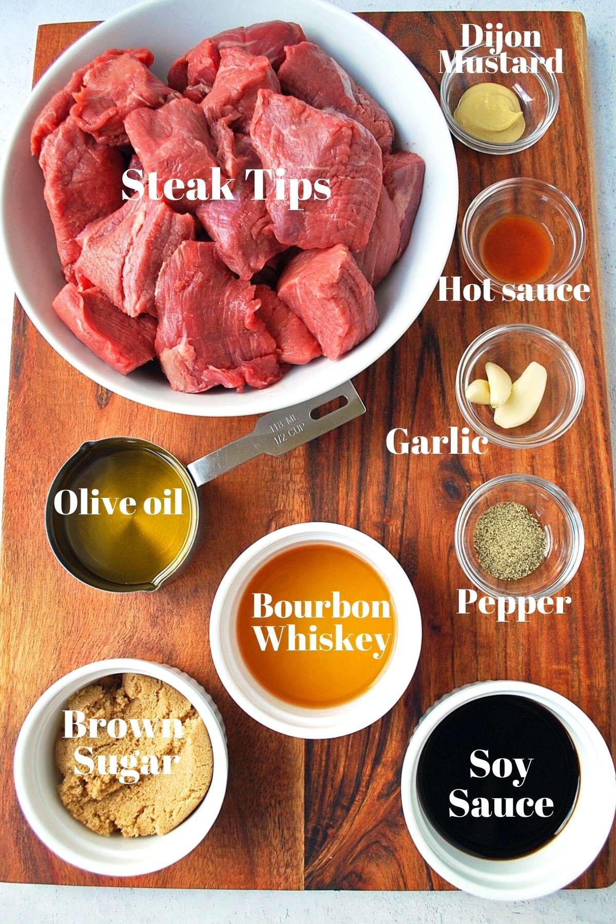 Steak tips, olive oil, brown sugar, whiskey, soy sauce,pepper, garlic, hot sauce, and dijon mustard on a cutting board