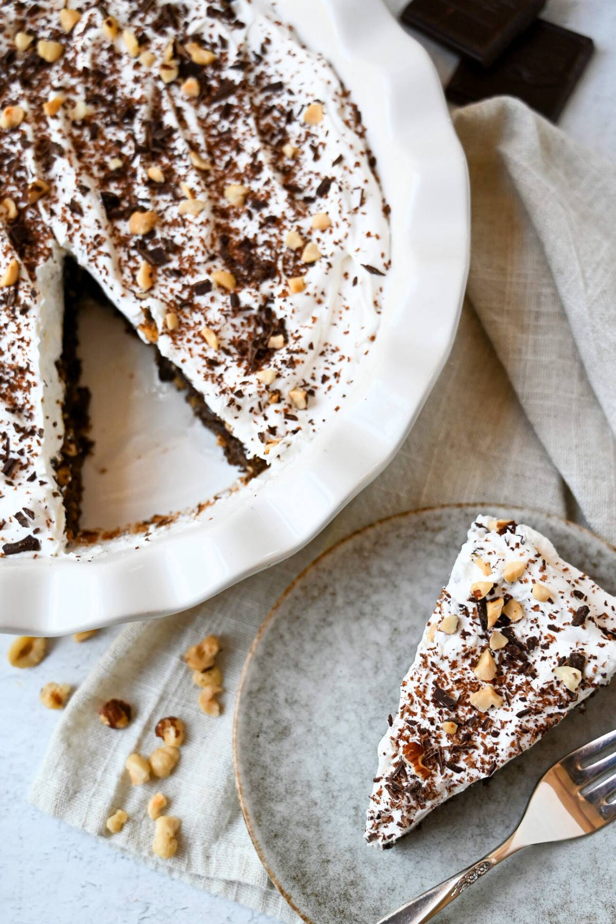 looking down at a chocolate hazelnut pie with a slice cut out served on a plate next to it and topped with chopped hazelnuts and grated chocolate