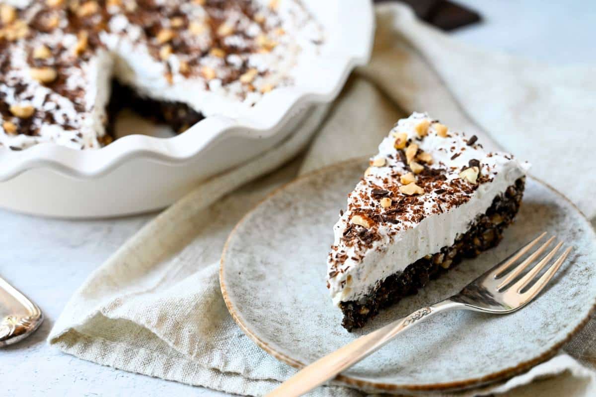 side view of a slice of chocolate hazelnut pie with cool whip and grated chocolate and hazelnuts on top on a plate with a fork with the sliced pie behind it