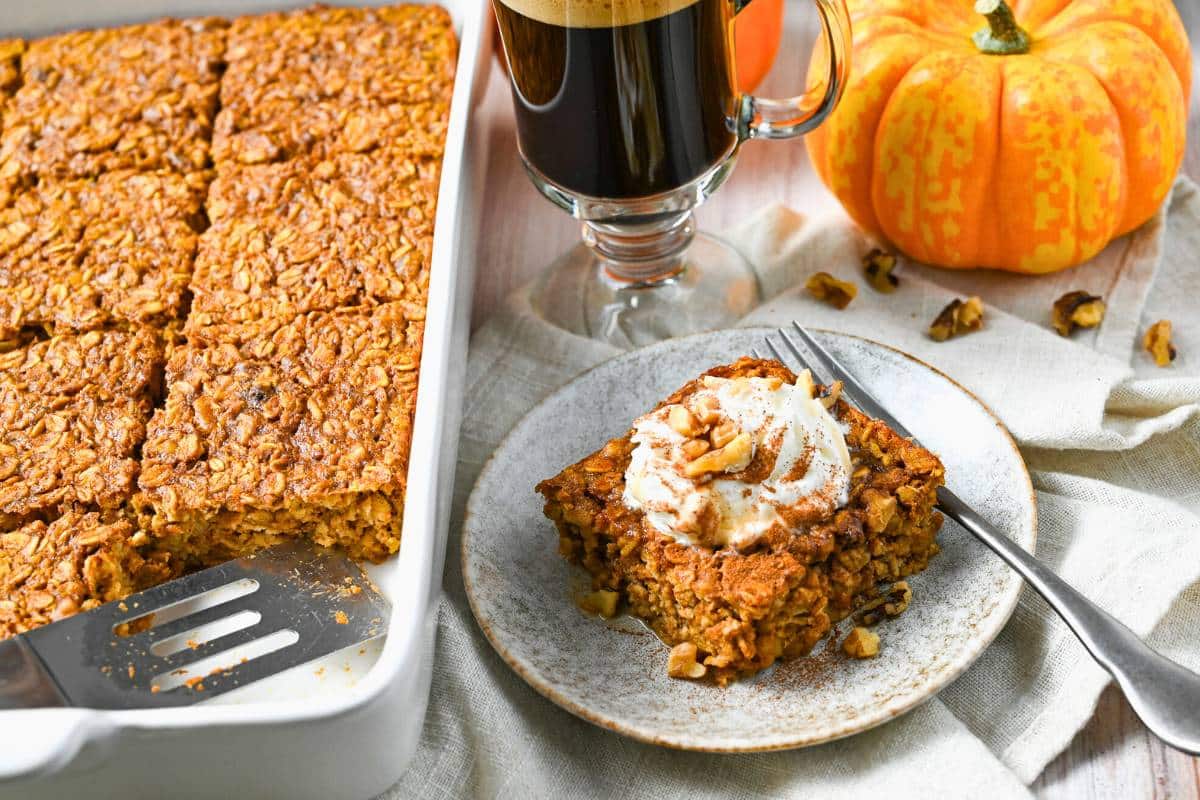 a slice of pumpkin baked oats on a plate with whipped cream, cinnamon, and walnuts with a fork and a pan of baked oats next to it