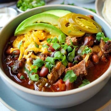close up photo of a bowl of brisket chili topped with jalapenos, avocado, scallions, and shredded cheese