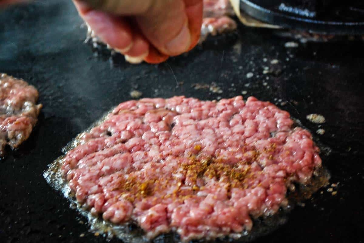 sprinkling burger seasoning on a smashed burger patty