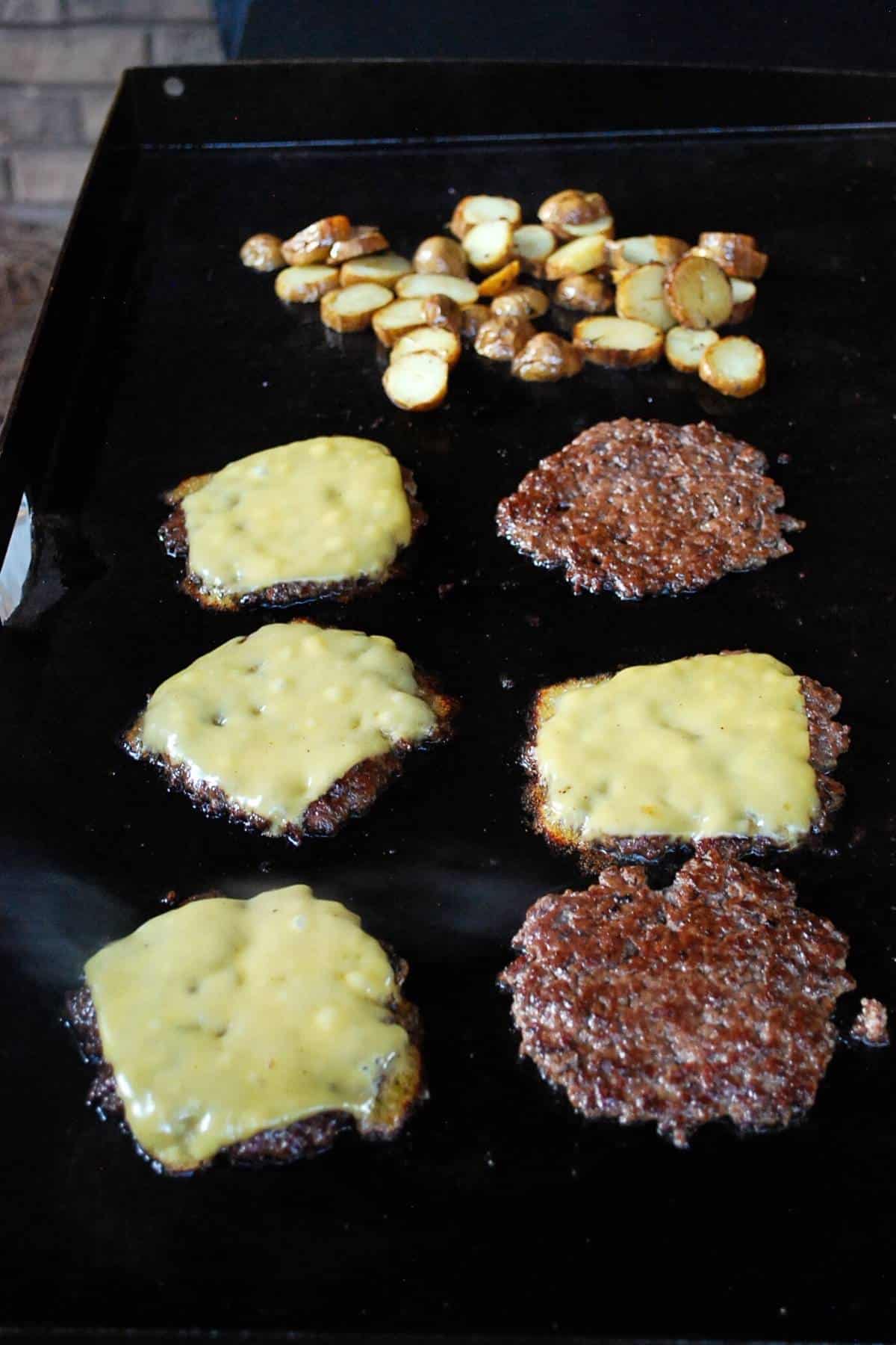 burger patties topped with cheese and potatoes cooking in the background