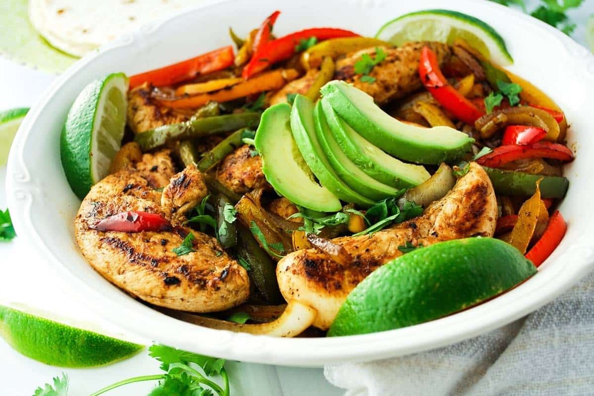 chicken fajitas in a large white serving bowl topped with sliced avocado and lime wedges