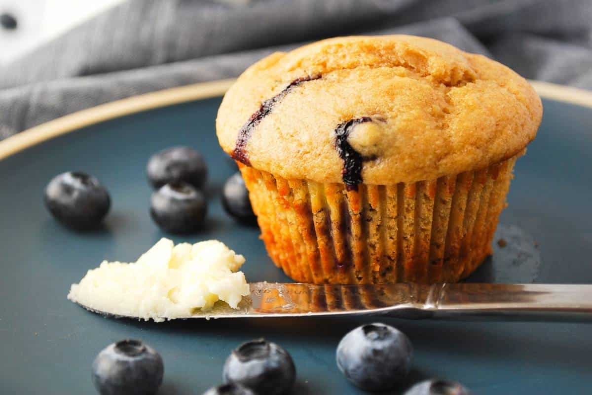 a blueberry muffin on a blue plate with a butter knife and fresh berries
