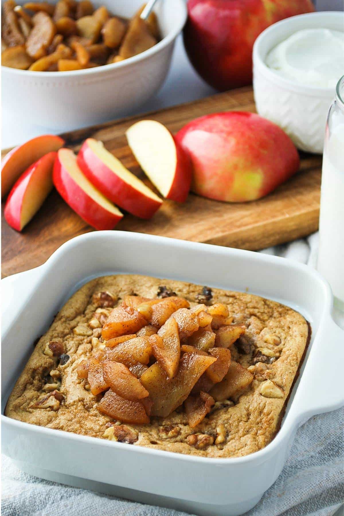 a pan of apple pie baked oats with cinnamon apples, a glass of milk, and sliced apples on a cutting board