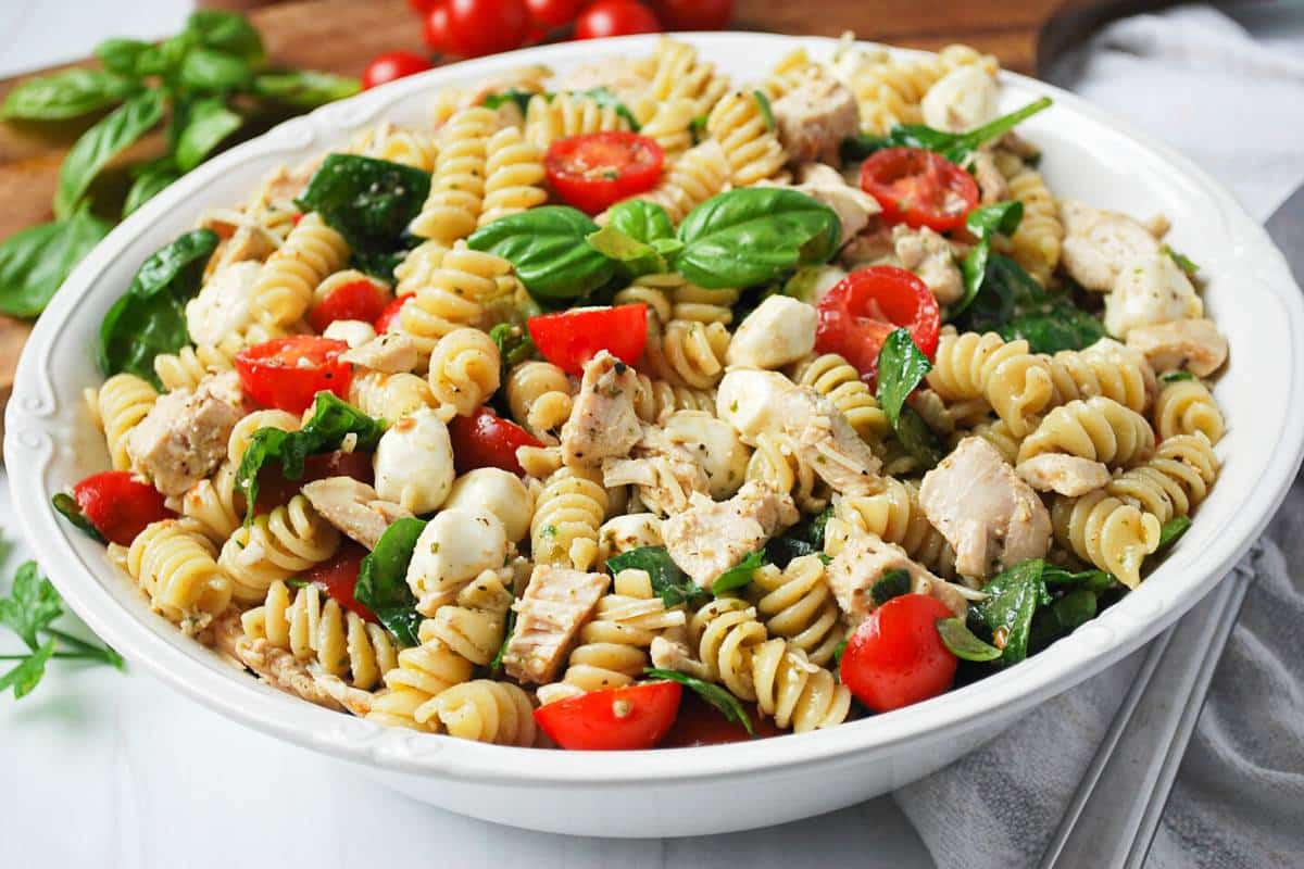 side view of a serving bowl of chicken pesto pasta salad with tomatoes, mozzarella, basil, spinach, and parmesan cheese