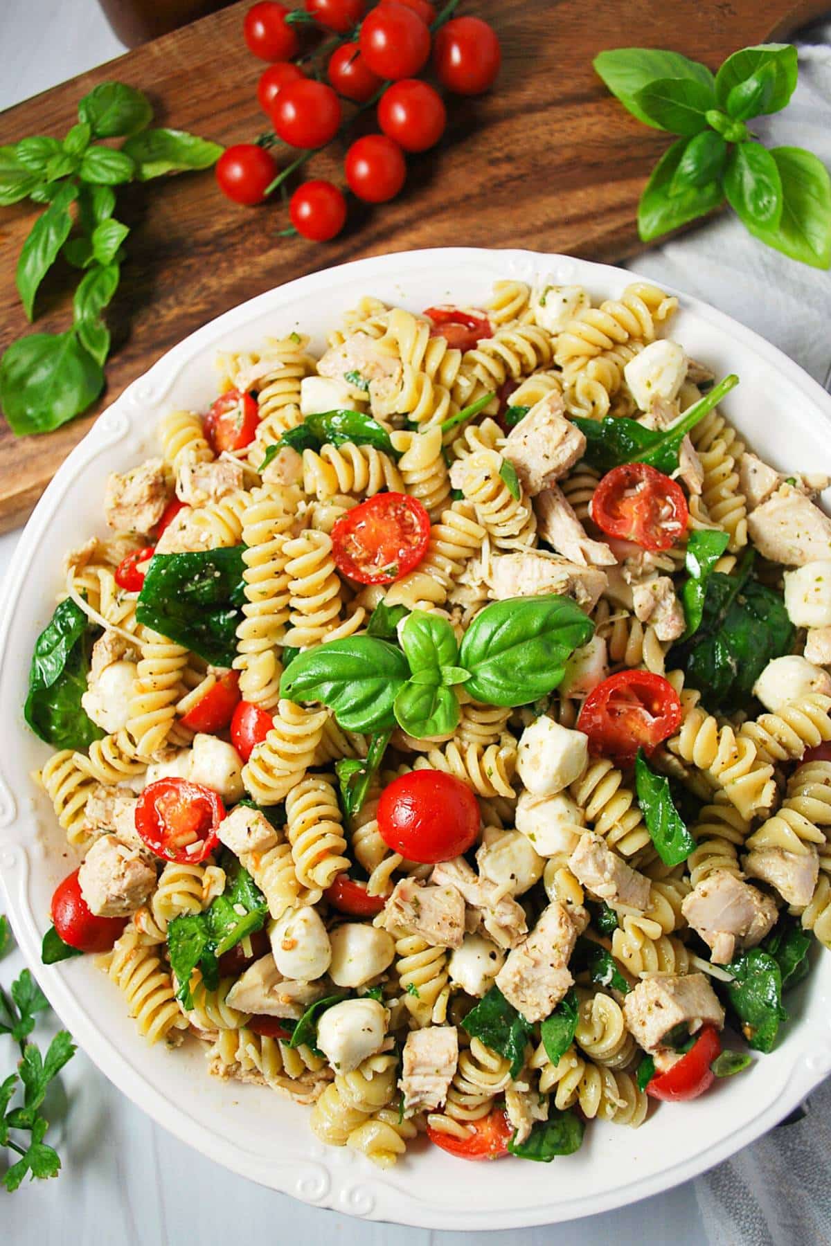 a serving bowl filled with pasta salad with a cutting board with cherry tomatoes and fresh basil in the background
