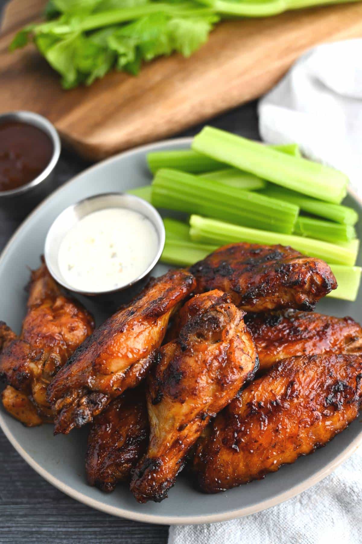 BBQ chicken wings on a plate with celery and a dish of ranch with a cutting board and napkin beside it