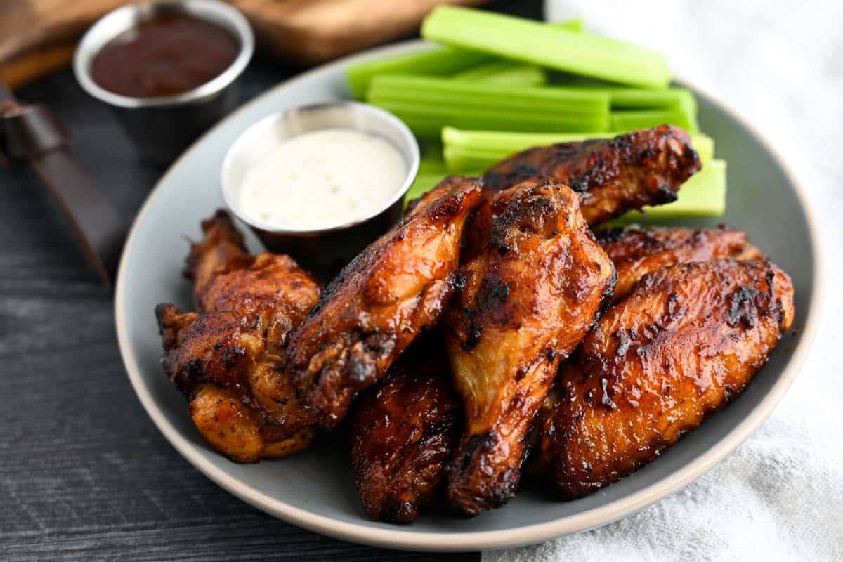 side view of a plate of wings with a dish of BBQ sauce and a cutting board with celery behind it