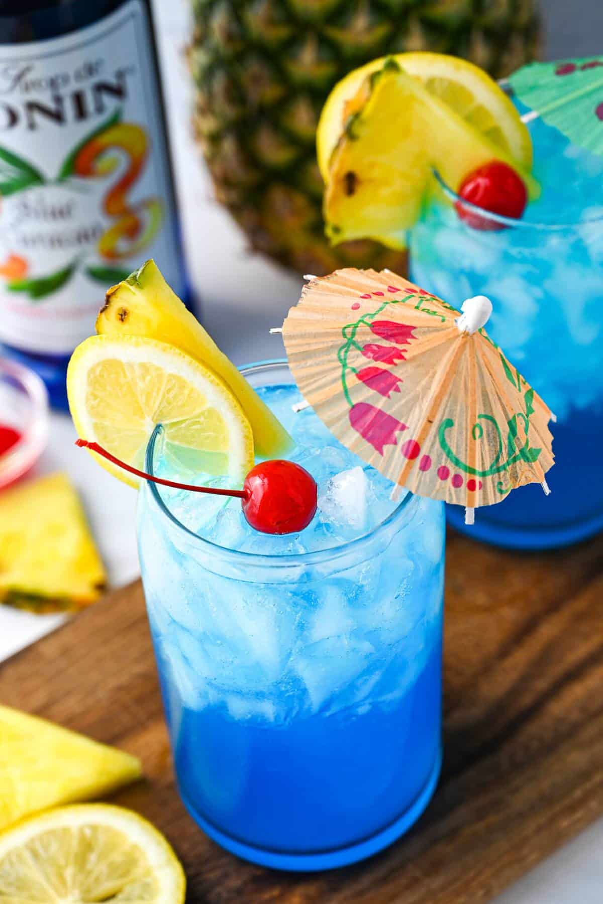 two glasses of non-alcoholic blue lagoon drink with cherries, little umbrellas, and fresh fruit with a pineapple behind them