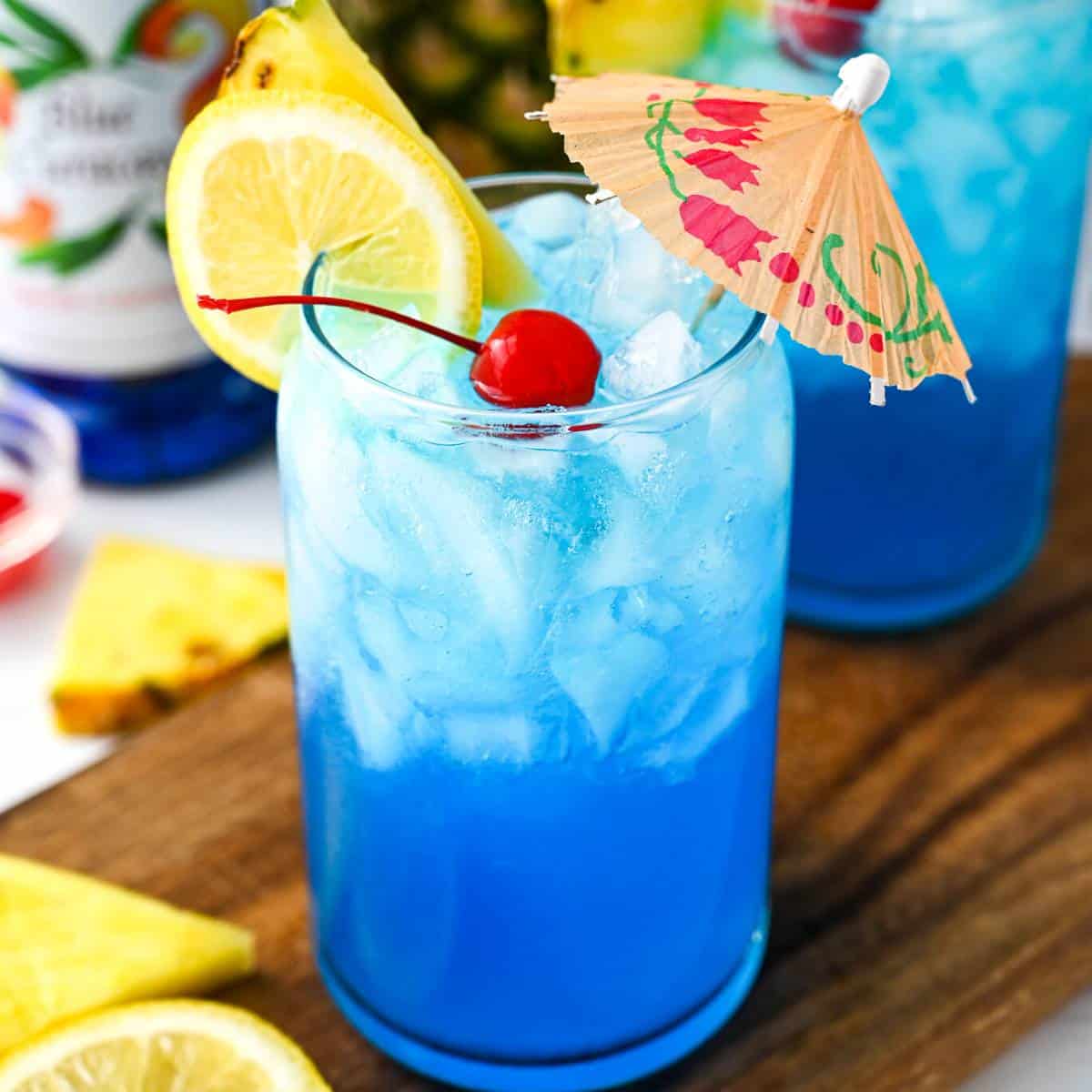 a close up of a blue lagoon mocktail in a glass with an umbrella, cherry, and pineapple and lemon slice