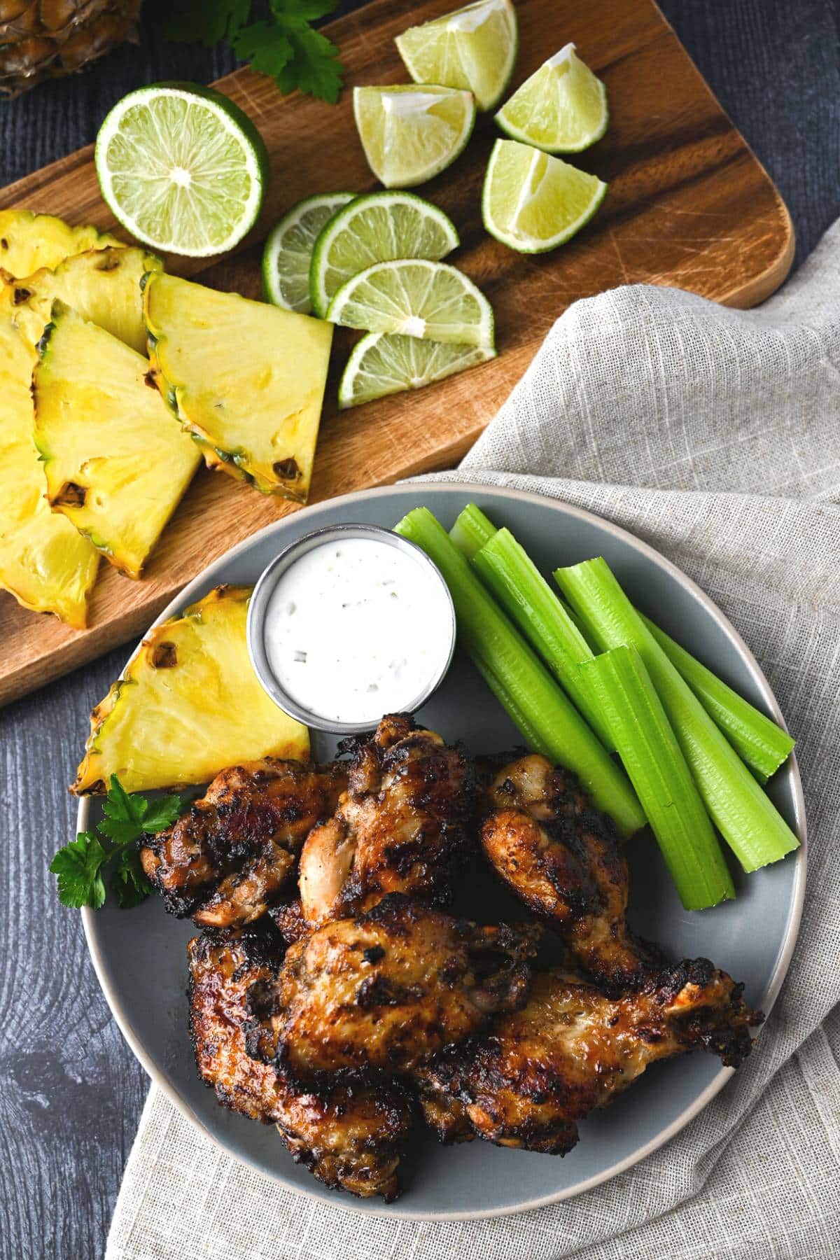 overhead photo of a plate of jerk wings with ranch, celery, and pineapple with a napkin and cutting board with pineapple and fresh lime