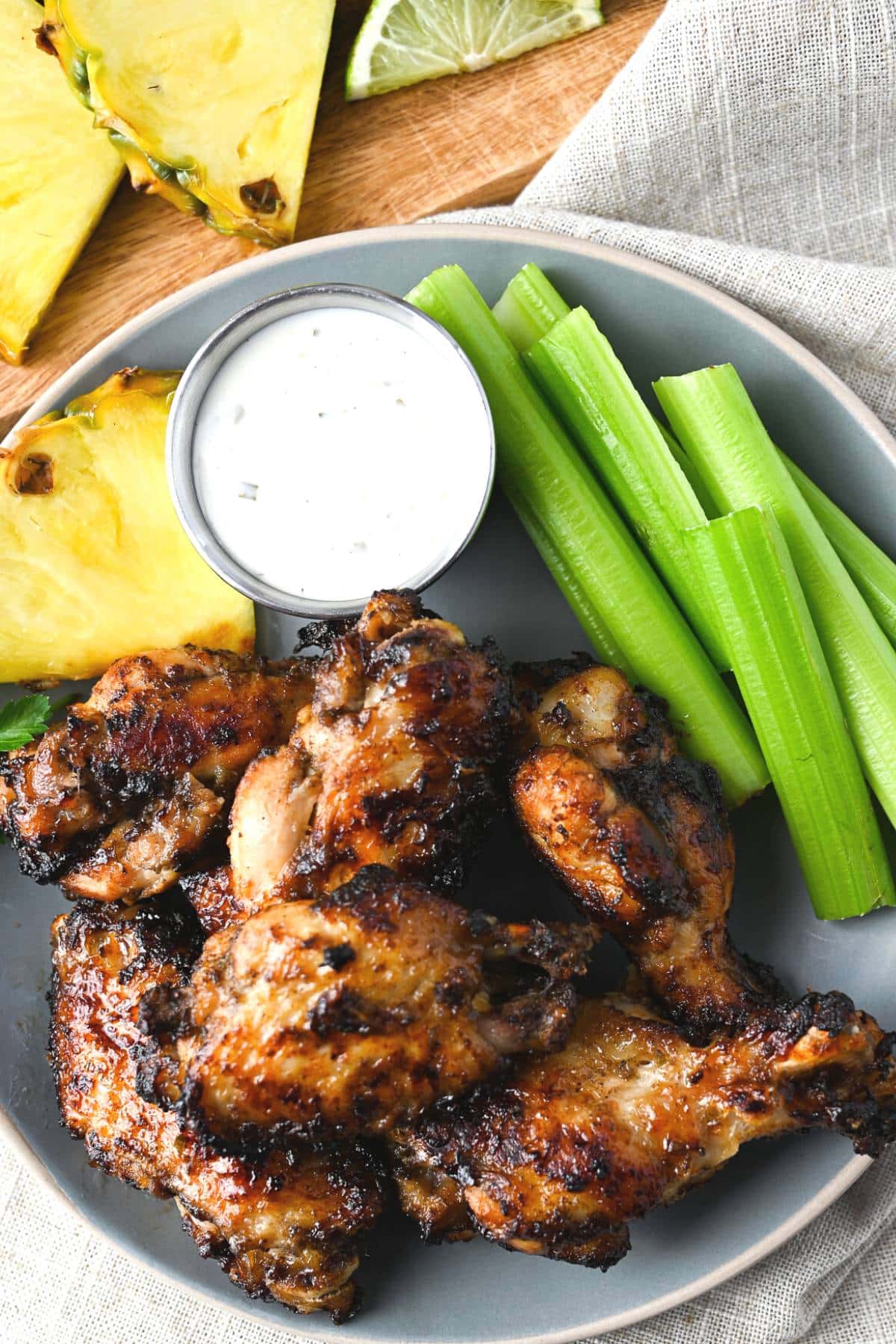 Jerk wings on a plate with celery, ranch, and a slice of pineapple with pineapple and lime in the background