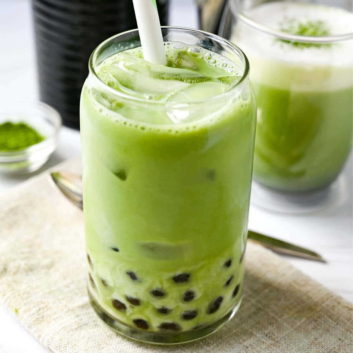 close up of a glass of iced matcha with boba with a mug of hot matcha behind it