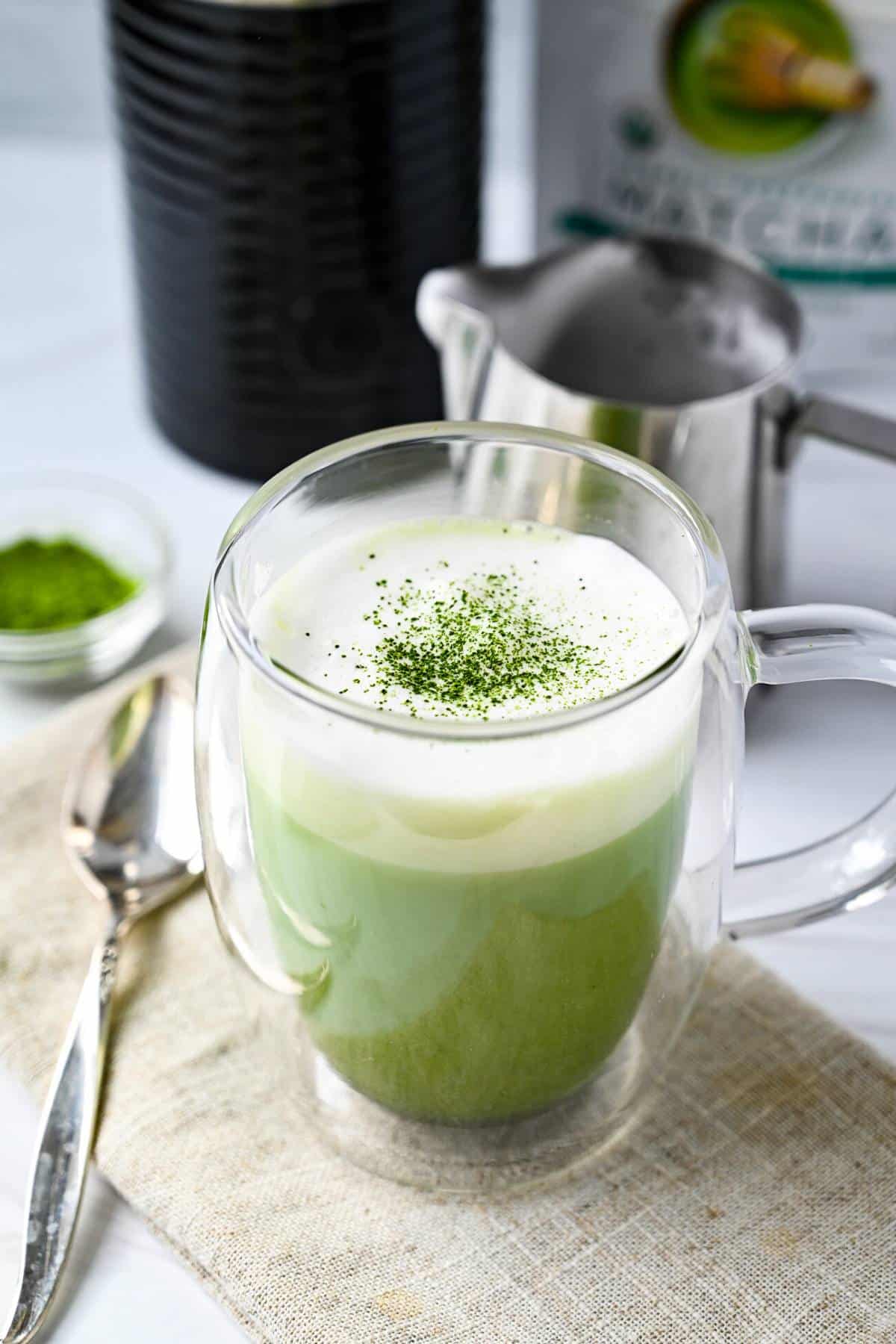 a matcha latte in a clear glass mug with froth and a sprinkle of matcha powder on top