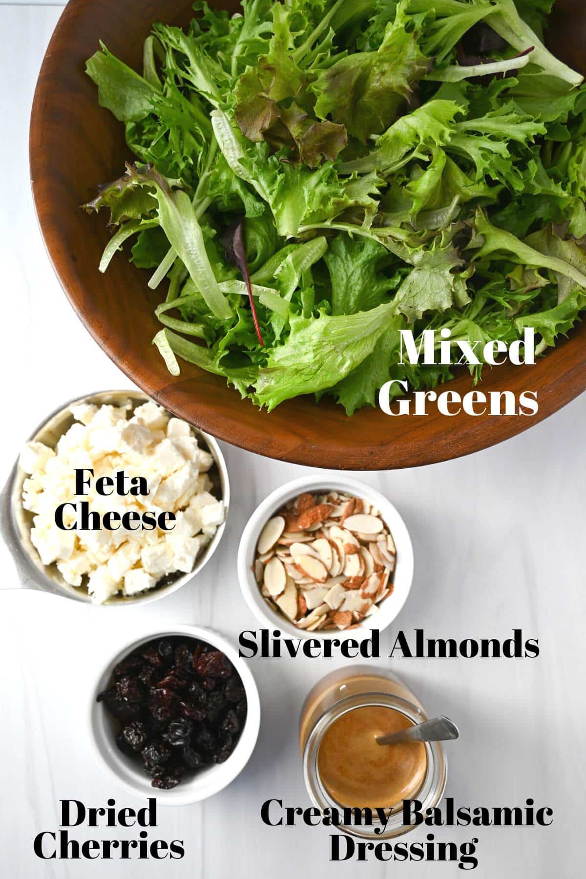 ingredients for house salad on a counter top: greens, dried cherries, feta, almonds and creamy balsamic dressing