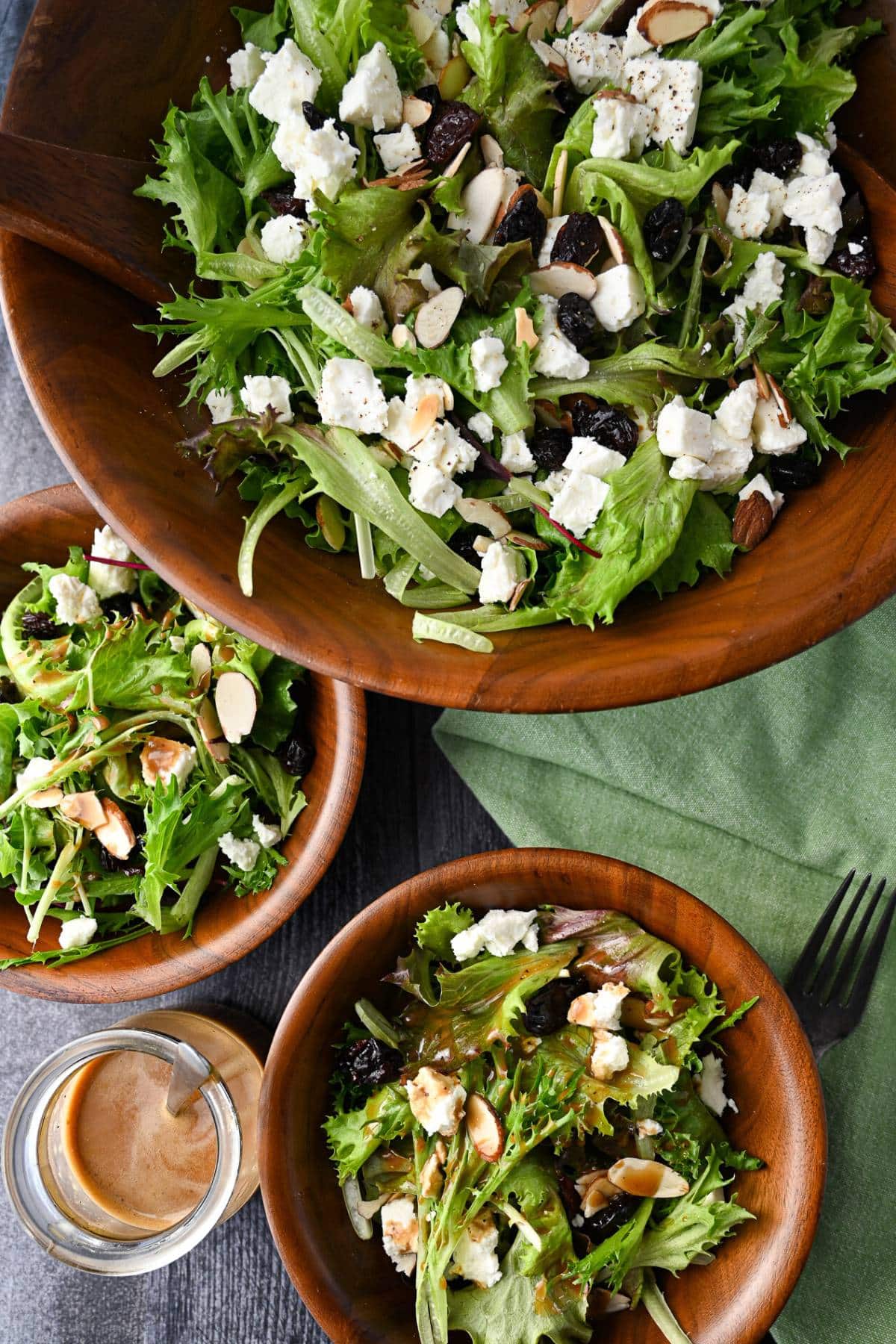 overhead photo of a large wood serving bowl full of greens with cherries, feta, and almonds and two small wood bowls with salad next to a jar of dressing