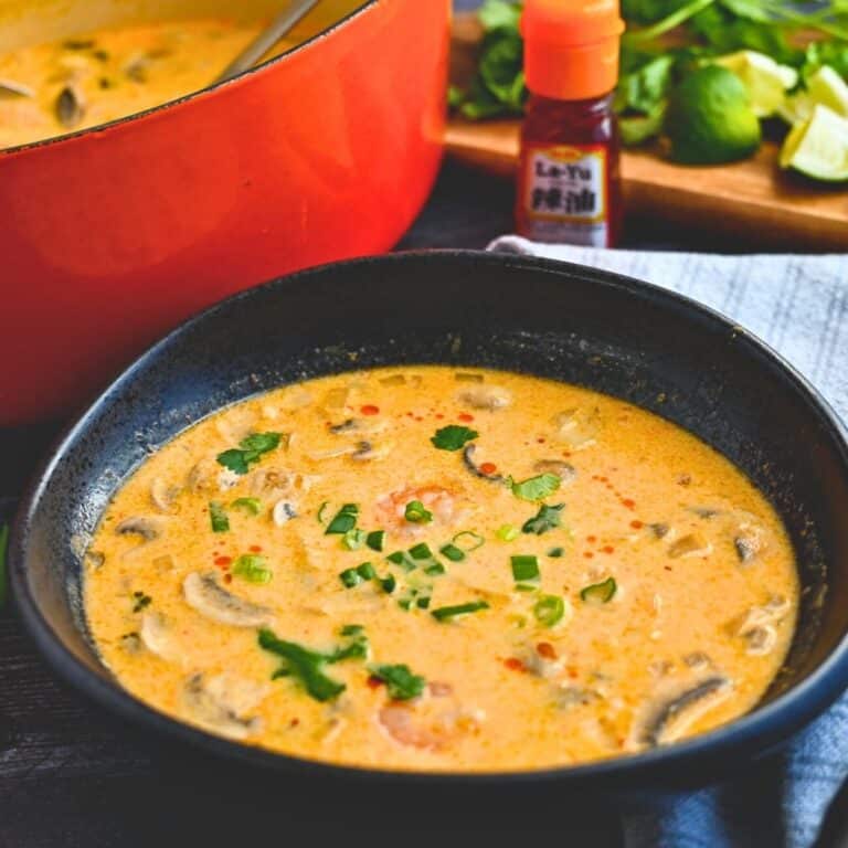 a bowl of Thai shrimp soup with a bottle of hot sauce, limes, and Dutch oven of soup behind it