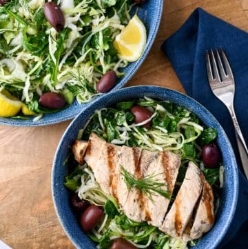 greek salad with cabbage, arugula, and grilled chicken in a blue bowl with a napkin and fork