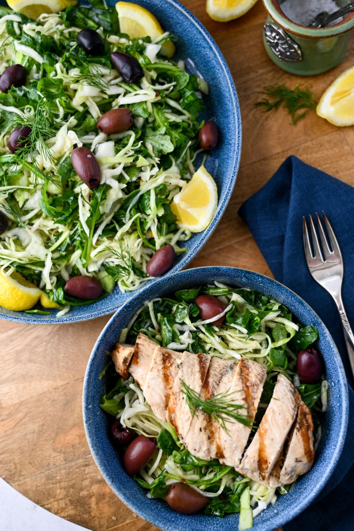 overhead photo of a bowl of Greek cabbage salad with olives and grilled chicken with a plate of cabbage salad and lemon wedges behind it