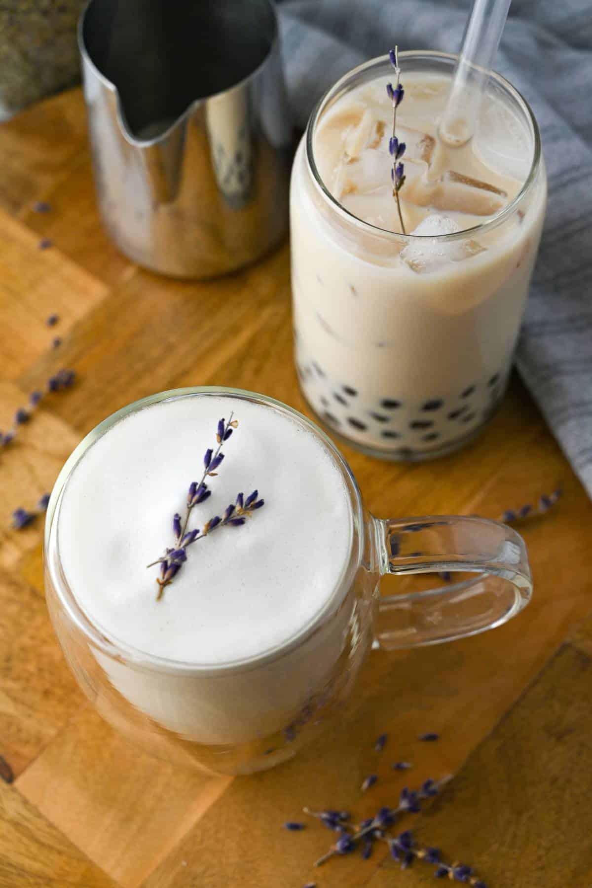 lavender milk tea in a mug with froth and a lavender stem with iced lavender boba tea in a glass behind it