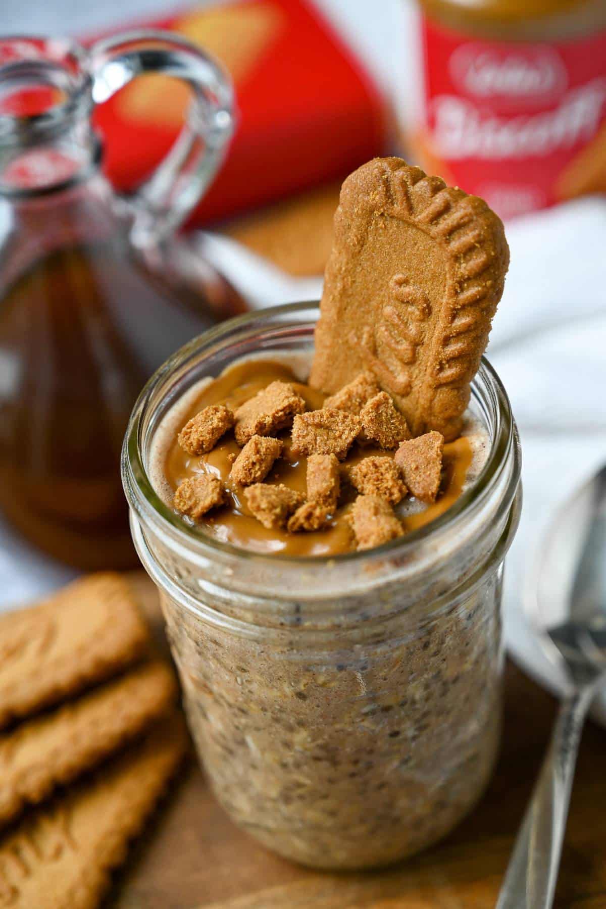 a jar of biscoff oats on a wood board with Biscoff cookies and cookie butter, syrup, and a package of Biscoff cookies in the background