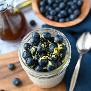 cheesecake overnight oats in a mason jar topped with fresh blueberries, lemon zest and maple syrup with a spoon, jar of syrup and bowl of blueberries next to it
