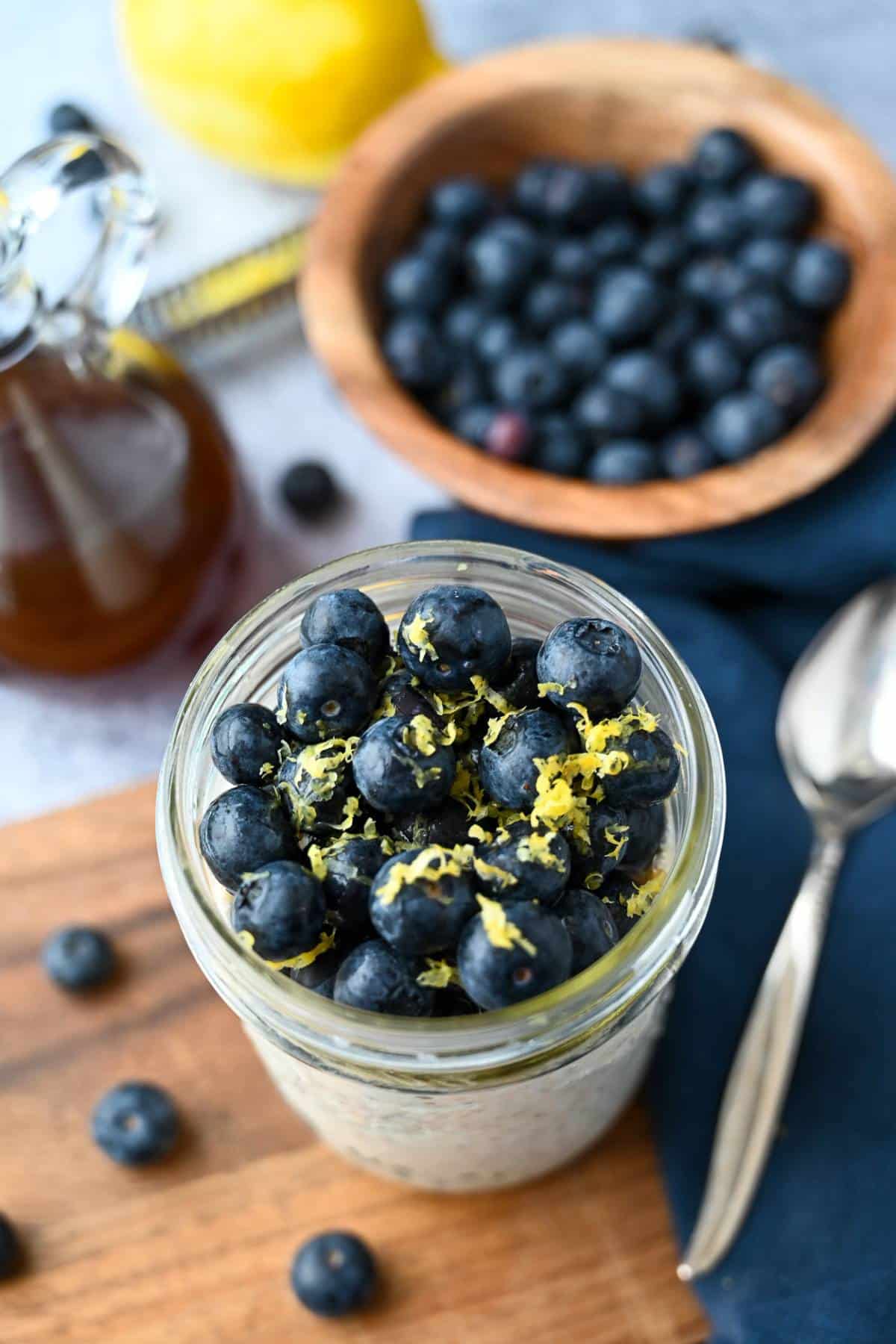 blueberry cheesecake oats in a jar with fresh blueberries and lemon zest with blueberries in a wooden bowl, a jar of syrup, and a lemon behind it