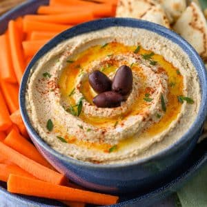 a bowl of high protein hummus with olive oil, parsley, and olives on top on a platter with carrots and pita bread
