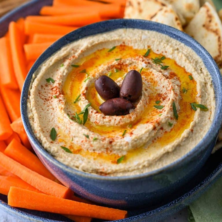 a bowl of high protein hummus with olive oil, parsley, and olives on top on a platter with carrots and pita bread