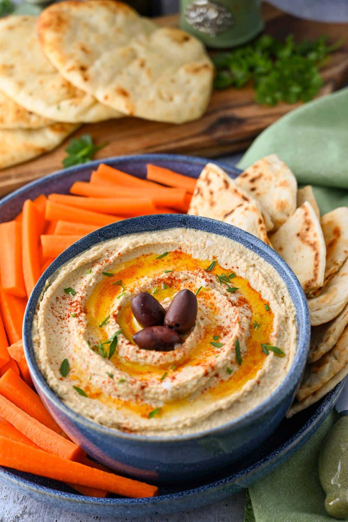 a bowl of high protein hummus with olives on top with pita bread and carrot sticks around it