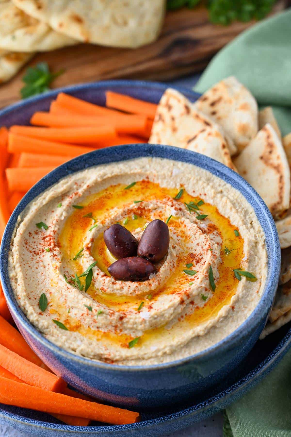 a platter of protein packed hummus with carrots and pita bread with a cutting board with bread and a cloth napkin behind it