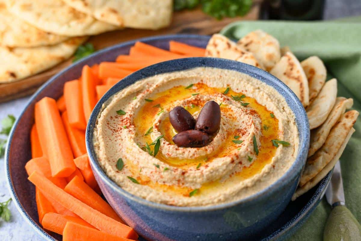 a bowl of hummus garnished with evoo, parsley, and kalamata olives with naan bread, pita slices, and carrots