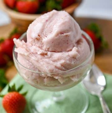 close up of a bowl of strawberry ice cream on top of a green napkin with a spoon and whole strawberries around it