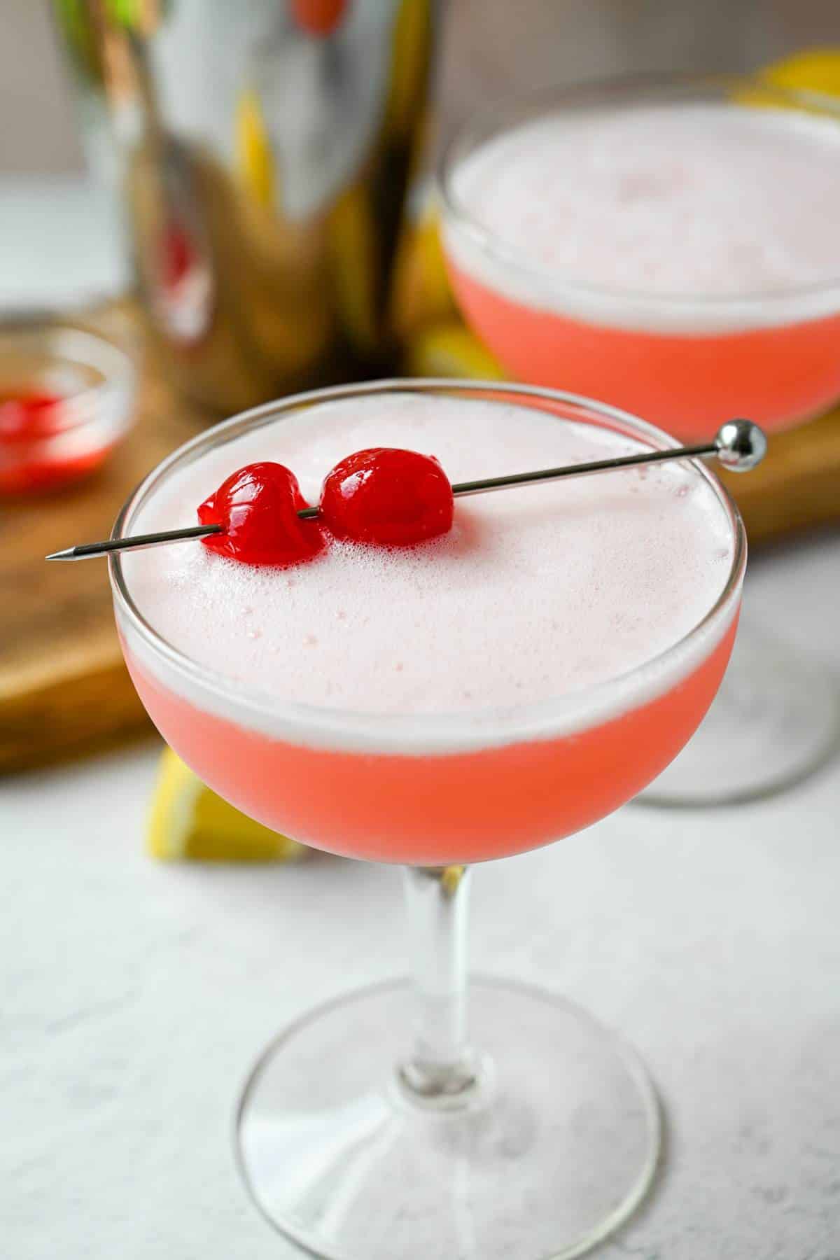two pink drink mocktails with a shaker and lemon slices and cherries on top