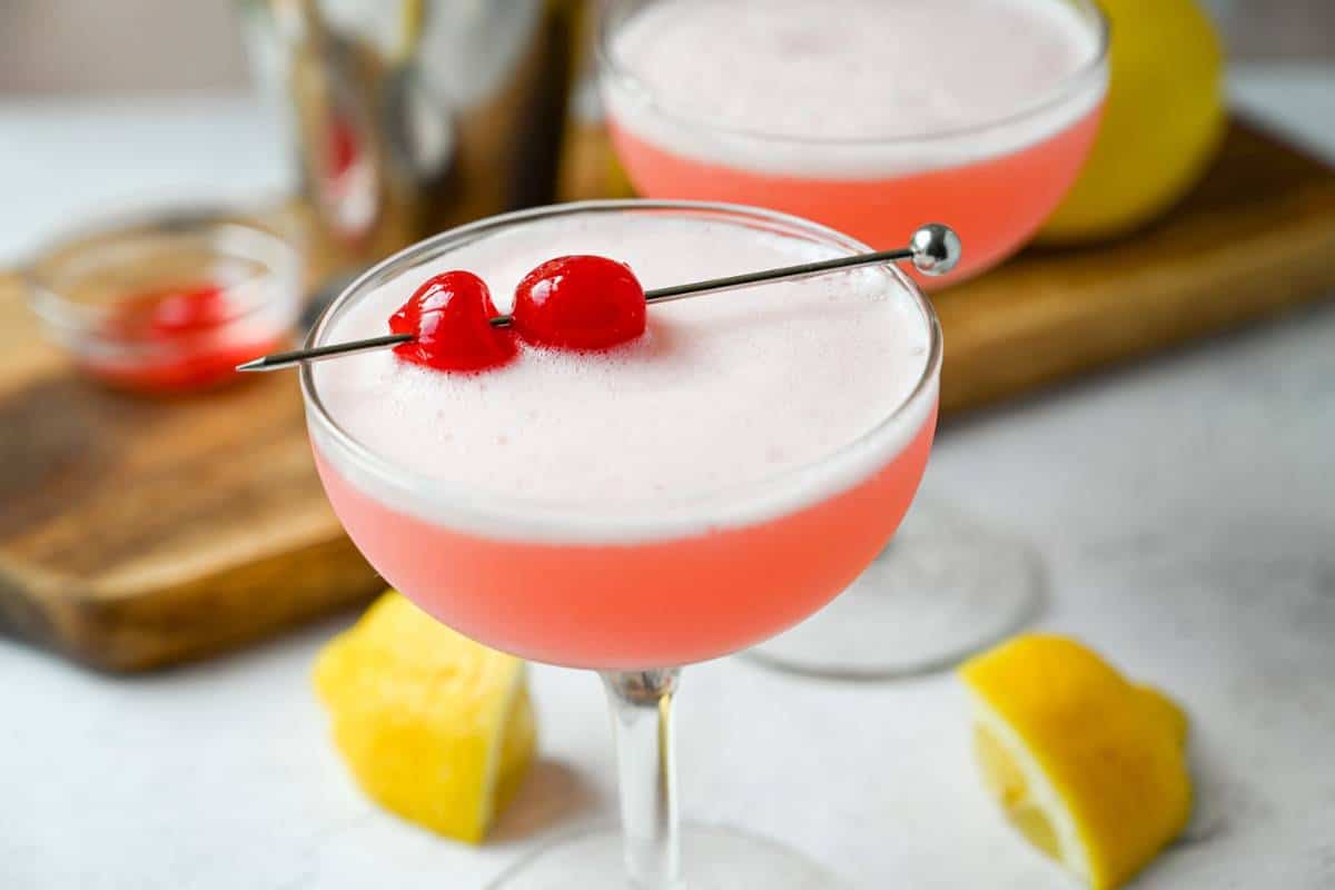 two pink lady mocktails in a clear glass with lemon slices next to them and cherries on top