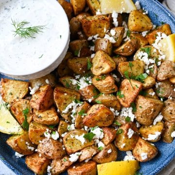 overhead photo of a platter of roasted baby potatoes with feta, herbs, lemon wedges and tzatziki sauce