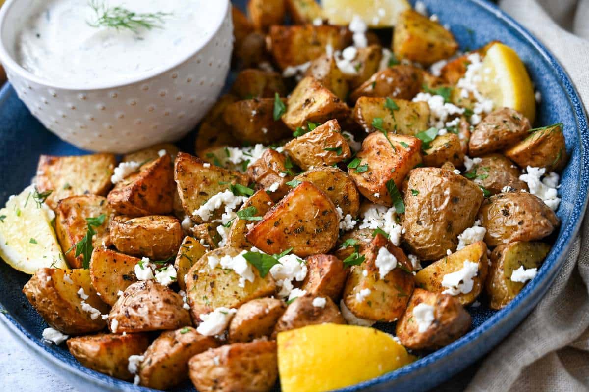 a platter of roasted greek potatoes garnished with herbs, lemon, feta, and a bowl of tzatziki