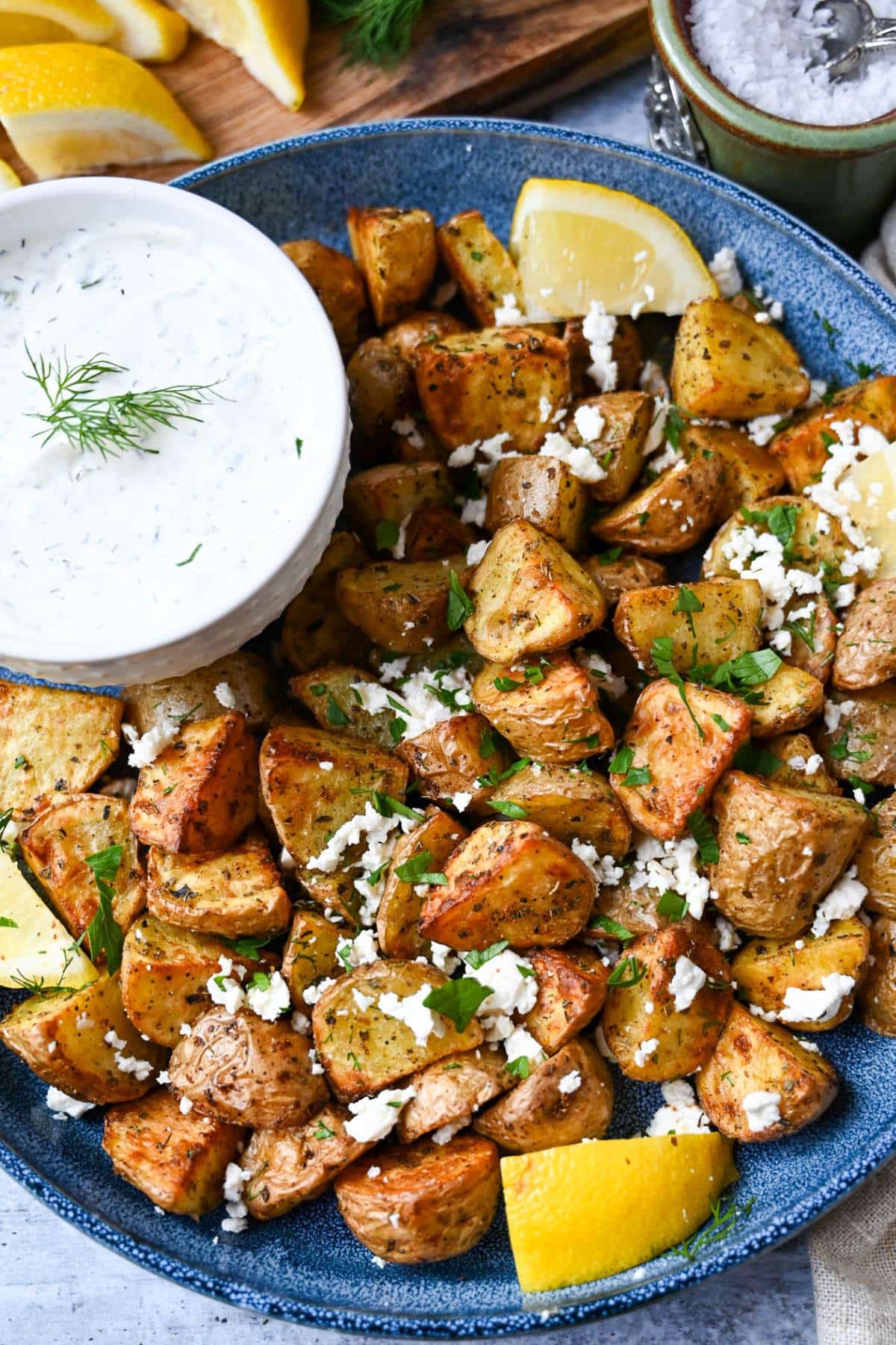 overhead photo of roasted potatoes on a blue platter served with lemon wedges, feta, and fresh herbs and a bowl of tzatziki sauce
